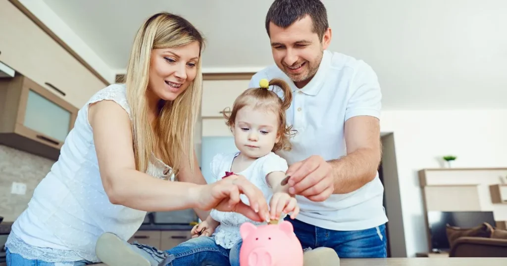 Mladá rodina s dcerou, která hází minci do růžového prasátka jako symbol spoření na budoucnost a penzi